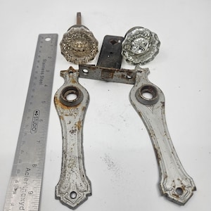 Peut inclure: Un ensemble comprenant deux poignées de porte en verre transparent à facettes, deux plaques de propreté métalliques argentées avec des traces de rouille, et un mécanisme de loquet central. Une règle en acier inoxydable indique 23 cm.