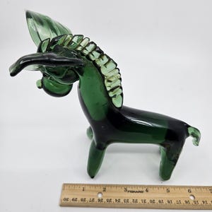 May include: A green glass horse figurine. The horse is stylized with a long neck, a flowing mane, and a curved tail. The glass is a deep shade of green, with some areas appearing darker. The figurine is approximately 6 inches long.