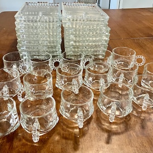 May include: A collection of clear glass tableware consisting of two stacks of rectangular trays with scalloped edges and multiple glass cups featuring round beaded handles. The glassware is transparent and arranged on a flat surface.
