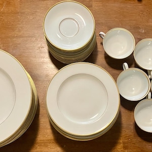 May include: A dinnerware set consisting of a stack of large white plates with gold rims and a subtle floral accent, a stack of smaller white saucers with gold rims, and eight individual white teacups featuring matching gold-trimmed edges.
