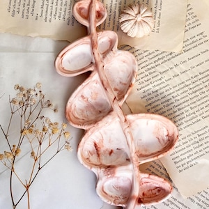 May include: A ceramic tray shaped like a eucalyptus branch with a marbled white and reddish-brown finish. The tray features multiple leaf-shaped indentations. A small, round, ribbed ceramic object and a sprig of dried baby's breath are nearby.