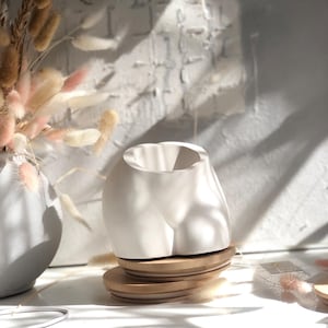 May include: A white ceramic planter shaped like a human torso, featuring a smooth matte finish and a wide circular opening at the top. The planter is positioned on top of two stacked circular bamboo lids, creating a stable base for the object.