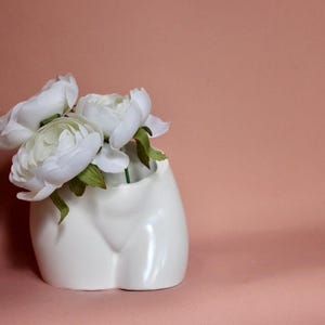 May include: A white ceramic vase shaped like a human torso, featuring a smooth, glossy finish. The vase contains a cluster of three white artificial flowers with green stems and leaves, positioned within the opening at the top of the torso form.