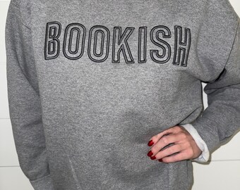 Puede incluir: Una sudadera gris con la palabra "BOOKISH" impresa en letras negras con contorno.