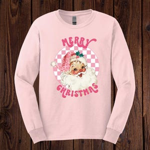 May include: Pink long-sleeve shirt with a retro-style illustration of Santa Claus with the text "Merry Christmas" in pink letters.