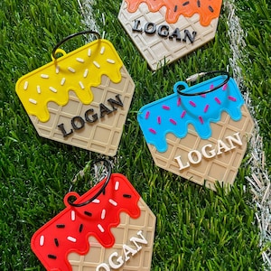 May include: Four ice cream cone-shaped luggage tags with the name "LOGAN" in white block letters. Each tag has a different color of "ice cream" and sprinkles: yellow, orange, blue, and red. The tags are on a green surface.