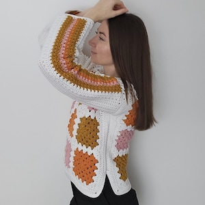 Könnte beinhalten: Ein weißer Häkelpullover mit bunten quadratischen Mustern in Orange-, Rosa- und Goldtönen. Die Ärmel haben gestreifte Akzente in ähnlichen Farben. Der Pullover wird mit einer schwarzen Hose kombiniert.