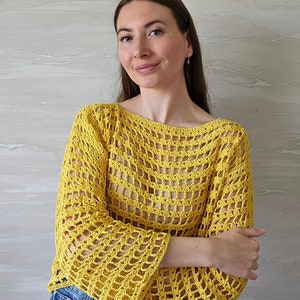 May include: A yellow crochet top with a loose, open-weave design. The top has a wide, boat neckline and long, flowing sleeves. The model is wearing the top with blue jeans.