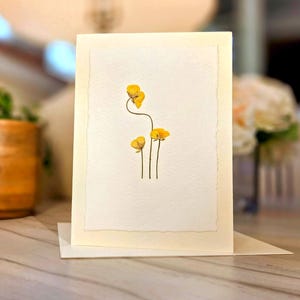 May include: A handmade greeting card featuring pressed yellow flowers on a cream-colored paper. The card is displayed on a white surface, with a blurred background including a plant in a terracotta pot and a vase of flowers.