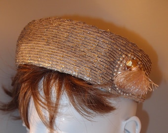 May include: A gold-toned, woven hat with a rounded crown, adorned with pearls and a small tassel. The hat is displayed on a white mannequin head with red lipstick and a pair of copper-colored earrings. A vintage-inspired accessory.