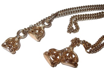 May include: A gold chain necklace with two ornate, decorative pendants. The chain is double-stranded and has a delicate, linked design. The pendants are intricate and feature a floral motif.