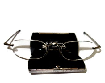 May include: Rimless eyeglasses with a silver-tone metal frame and clear lenses rest on a black eyeglass case. The case has a silver clasp and a black interior. The glasses are open.