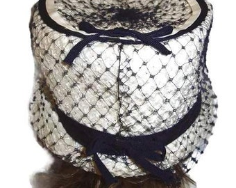 May include: A white hat with a black net design and a black ribbon. The hat is made of straw and has a wide brim.