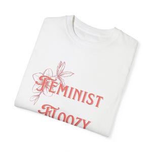 Puede incluir: Camiseta blanca con un diseño floral rosa y el texto "Feminist Floozy" impreso en la parte delantera.