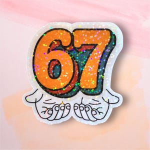 May include: A glittery sticker featuring the number "67" in orange with a teal outline, set against a white background. The number is stylized with a retro font and is held by two cartoon hands. The sticker has a holographic glitter effect.
