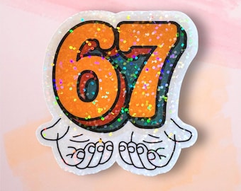 May include: A glittery sticker featuring the number "67" in orange with a teal outline, set against a white background. The number is stylized with a retro font and is held by two cartoon hands. The sticker has a holographic glitter effect.