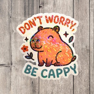This sticker features an adorable, cartoon-style capybara sitting contentedly with a big, peaceful smile and closed eyes. The capybara has a light brown body with darker brown accents for the ears, outline, and tiny fur markings.  Above the capybara, the text reads “DON’T WORRY,” in playful, rounded orange letters. Below the capybara, the phrase “BE CAPPY” is written in bold, teal-blue bubble letters — a clever pun combining “happy” and “capybara.”