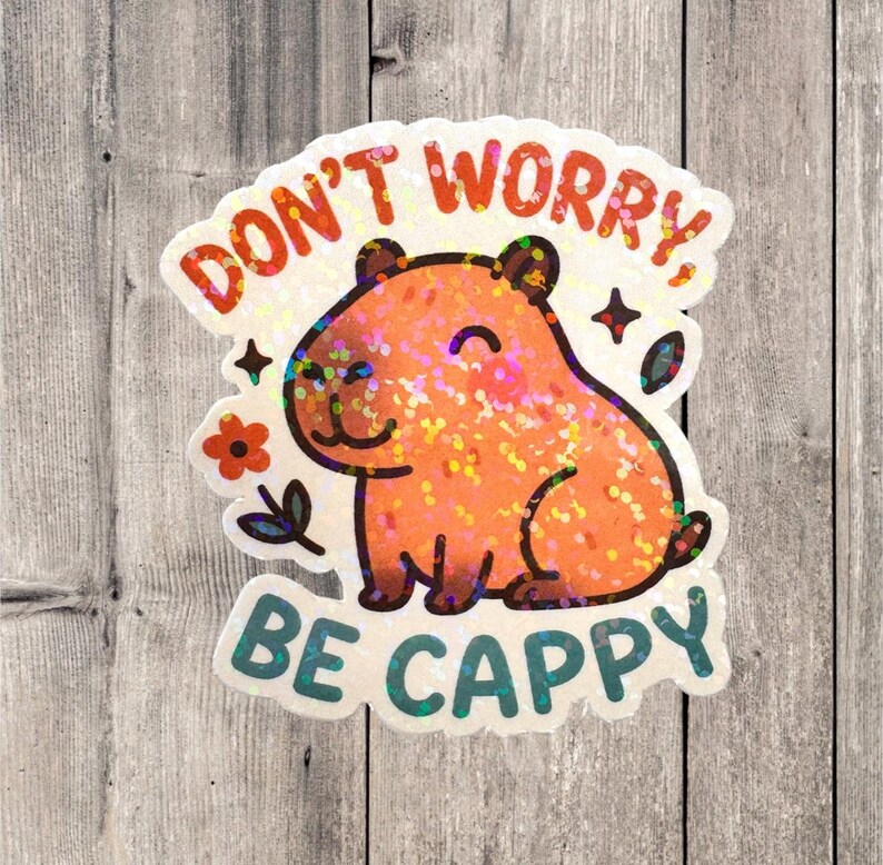 This sticker features an adorable, cartoon-style capybara sitting contentedly with a big, peaceful smile and closed eyes. The capybara has a light brown body with darker brown accents for the ears, outline, and tiny fur markings.  Above the capybara, the text reads “DON’T WORRY,” in playful, rounded orange letters. Below the capybara, the phrase “BE CAPPY” is written in bold, teal-blue bubble letters — a clever pun combining “happy” and “capybara.”