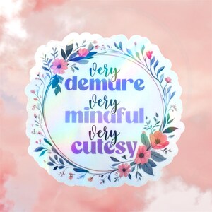A circular holographic sticker with a scalloped edge, featuring a delicate floral wreath in pink and blue tones. The text "very demure very mindful very cutesy" is written in a mix of elegant script and bold purple/blue lettering. The entire sticker has a smooth, pearlescent holographic sheen that reflects a rainbow spectrum.