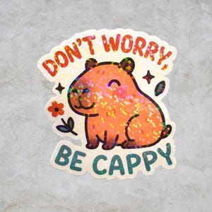 This sticker features an adorable, cartoon-style capybara sitting contentedly with a big, peaceful smile and closed eyes. The capybara has a light brown body with darker brown accents for the ears, outline, and tiny fur markings.  Above the capybara, the text reads “DON’T WORRY,” in playful, rounded orange letters. Below the capybara, the phrase “BE CAPPY” is written in bold, teal-blue bubble letters — a clever pun combining “happy” and “capybara.”