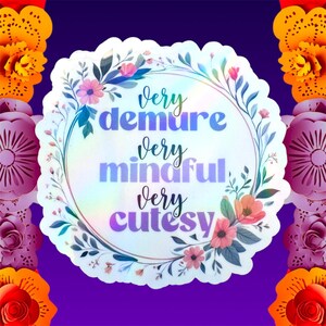 A circular holographic sticker with a scalloped edge, featuring a delicate floral wreath in pink and blue tones. The text "very demure very mindful very cutesy" is written in a mix of elegant script and bold purple/blue lettering. The entire sticker has a smooth, pearlescent holographic sheen that reflects a rainbow spectrum.