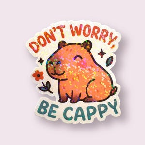 This sticker features an adorable, cartoon-style capybara sitting contentedly with a big, peaceful smile and closed eyes. The capybara has a light brown body with darker brown accents for the ears, outline, and tiny fur markings.  Above the capybara, the text reads “DON’T WORRY,” in playful, rounded orange letters. Below the capybara, the phrase “BE CAPPY” is written in bold, teal-blue bubble letters — a clever pun combining “happy” and “capybara.”