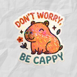 This sticker features an adorable, cartoon-style capybara sitting contentedly with a big, peaceful smile and closed eyes. The capybara has a light brown body with darker brown accents for the ears, outline, and tiny fur markings.  Above the capybara, the text reads “DON’T WORRY,” in playful, rounded orange letters. Below the capybara, the phrase “BE CAPPY” is written in bold, teal-blue bubble letters — a clever pun combining “happy” and “capybara.”