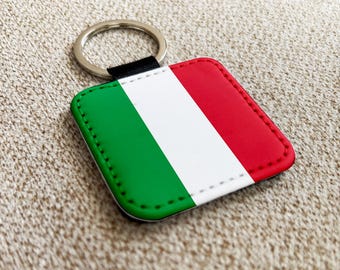May include: Square keychain featuring the Italian flag design. The keychain has a red, white, and green color scheme, with a metal ring for attaching keys. The edges are stitched with green thread.