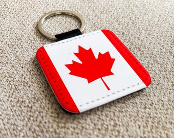 May include: A square keychain featuring the Canadian flag. The design includes a red maple leaf on a white background, framed by a red border. The keychain has a silver metal ring and a black fabric loop.