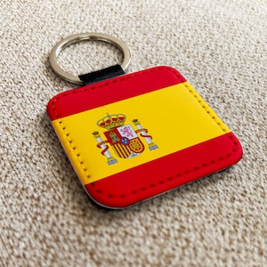 Op de afbeelding: Een vierkante sleutelhanger met het ontwerp van de Spaanse vlag. De sleutelhanger heeft een rode rand en een gele midden met het Spaanse wapen. Het bevat een zilveren sleutelring en een zwarte stoffen lus.