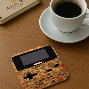 May include: A wooden coaster with a retro video game design featuring a Marvel comic book collage. The coaster has a black screen and control buttons. A white coffee cup with black coffee sits on a saucer nearby.