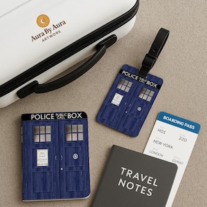 May include: A white suitcase with a black handle and the text "Aura By Aura ARTWORK". Includes a blue Police Box luggage tag, a Police Box notebook, a boarding pass, and a gray notebook with "TRAVEL NOTES" printed on it.