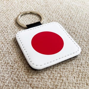 Może przedstawiać: Brelok do kluczy w kształcie kwadratu z flagą Japonii, z czerwonym kołem na białym tle. Brelok ma srebrny metalowy pierścień i czarny pasek. Całość umieszczona jest na teksturowanej, beżowej powierzchni.