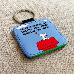 Puede incluir: Un llavero cuadrado con Snoopy sentado en su caseta roja sobre un fondo azul claro. El llavero tiene el texto "Keep looking up that is the secret of life" y un llavero plateado.