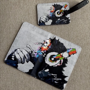 May include: Two grey luggage tags featuring a colourful illustration of a monkey wearing headphones. The monkey is depicted in a graffiti-style art form, with vibrant splashes of colour. One tag has a black strap.