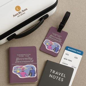 May include: A white suitcase with a black handle and the text "Aura By Aura Artwork". A purple passport holder, luggage tag, and notebook with the text "Handbook for the Recently Deceased" and "Travel Notes". A boarding pass is also visible.