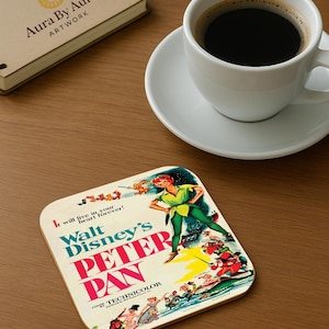 May include: A square coaster with a vintage Peter Pan movie poster design. The coaster has a cream background with the movie title in red and blue. A white coffee cup and saucer are on a wooden surface.