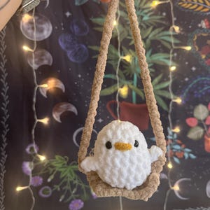May include: A white crocheted bird with a yellow beak sits in a brown crocheted swing. The swing is hanging from a beige rope.