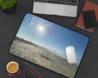 May include: A rectangular mousepad featuring a beach scene with a bright sun and blue sky. A white computer mouse sits on the right side of the pad. The pad is surrounded by a black border. A laptop, coffee cup, and writing utensils are also visible.