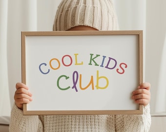 May include: A child wearing a white knitted hat holds a white framed sign that says "COOL KIDS CLUB" in colorful letters. The sign is held in front of the child's face. The child is sitting on a beige couch with pillows.