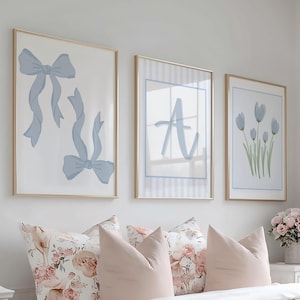 Könnte beinhalten: Ein Schlafzimmer mit einem Bett mit floralen Kissen und einer rosa Bettdecke. Über dem Bett hängen drei gerahmte Drucke mit blauen Schleifen-, Buchstaben- und Blumenmotiven. Auf der linken Seite befindet sich ein weißer Nachttisch mit einer Lampe.