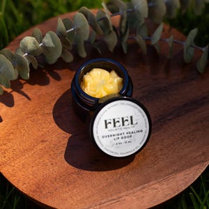 May include: A small, open black jar of yellow lip balm sits on a wooden plate, with the lid beside it. The lid displays the text "FEEL Holistic Health Overnight Healing Lip Goop". Eucalyptus leaves are in the background.