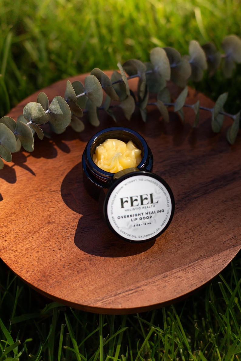 May include: A small, open black jar of yellow lip balm sits on a wooden plate, with the lid beside it. The lid displays the text "FEEL Holistic Health Overnight Healing Lip Goop". Eucalyptus leaves are in the background.
