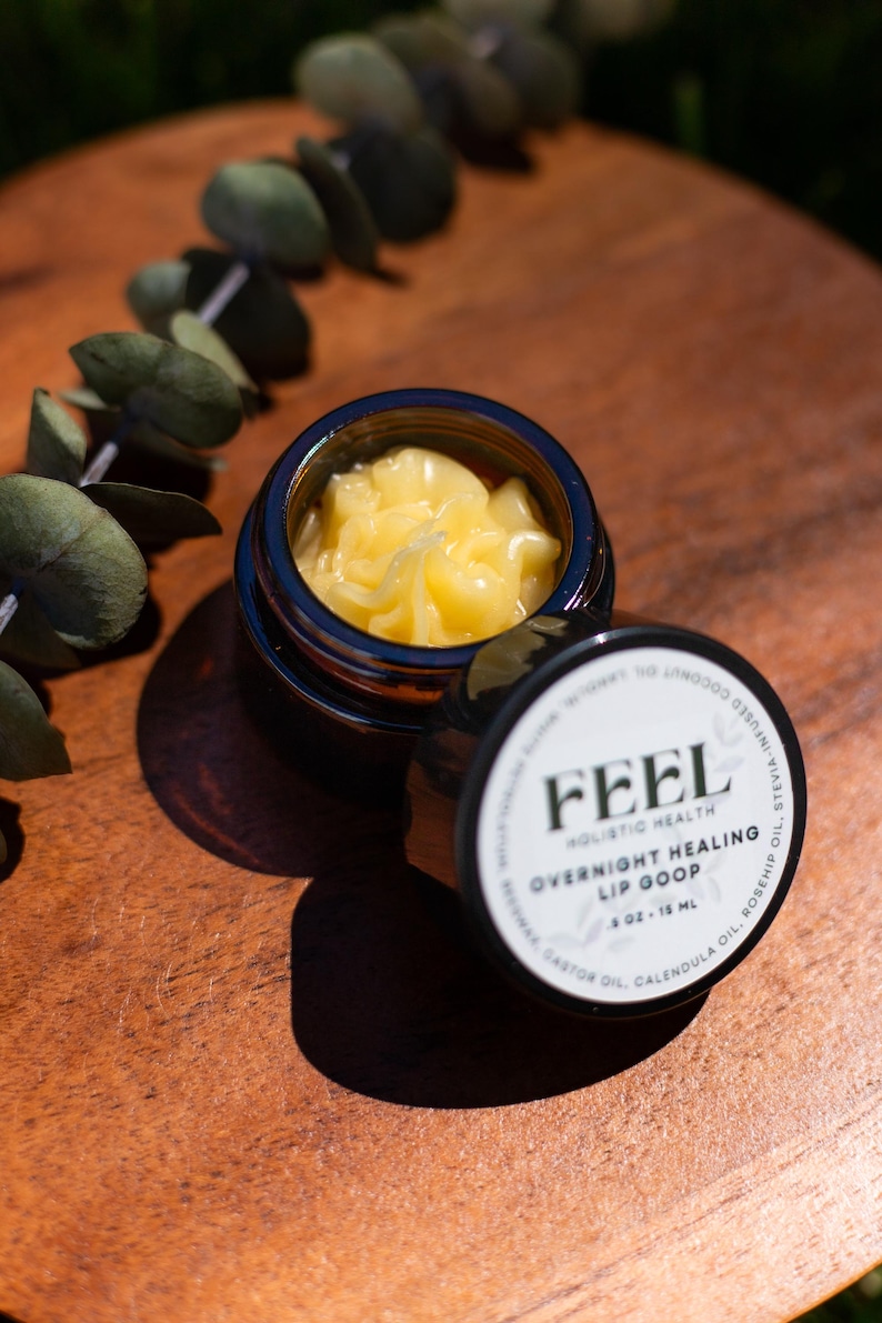 May include: A jar of "FEEL Holistic Health Overnight Healing Lip Goop" with a yellow balm inside. The jar is open, revealing the product, and the lid is next to it. The label includes the ingredients: castor oil, calendula oil, and rosehip oil. The jar contains 0.5 oz or 15 mL.