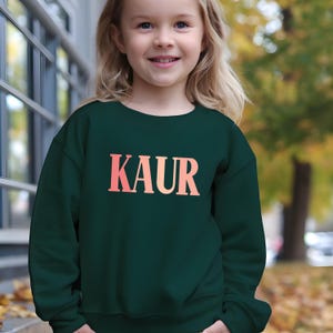 Peut inclure: Un sweat-shirt vert foncé avec le nom "KAUR" imprimé en dégradé rose et orange. Le sweat-shirt est porté par une jeune fille blonde souriante. L'arrière-plan est flou, avec un bâtiment et des arbres visibles.