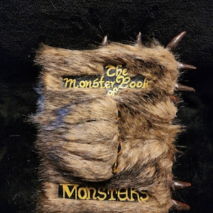 Może przedstawiać: Brązowa książka z imitacji futra z pozłacanymi napisami "The Monster Book of Monsters". Książka ma brązowe, kolczaste krawędzie.