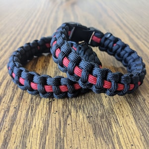 May include: Two black and red paracord bracelets with a black plastic buckle. The bracelets are woven with a red stripe running through the black paracord.