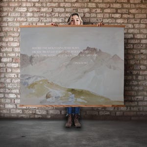 May include: A large rectangular canvas print with a watercolor mountain landscape in muted tones of beige, gray, and green. The print includes the text "BEFORE THE MOUNTAINS WERE BORN...FROM EVERLASTING TO EVERLASTING YOU ARE GOD - PSALM 90:2". The print is held up by a person.