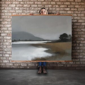 Puede incluir: Una gran pintura de paisaje con marco de madera. La obra representa una escena serena con una masa de agua, una montaña al fondo y un árbol a la derecha. La paleta de colores es apagada, con tonos grises, marrones y blancos.