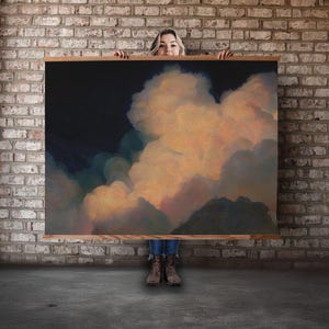 May include: A large rectangular canvas art piece featuring a dramatic cloudscape. The artwork showcases a dark blue sky contrasting with fluffy, peach-colored clouds. The canvas is framed with a light-colored wooden border.